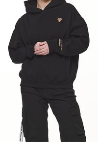 Persona que lleva puesto un sudadero negro con un pequeño logo de palmera y texto en la manga, combinado con pantalones cargo negros con bolsillos laterales.
