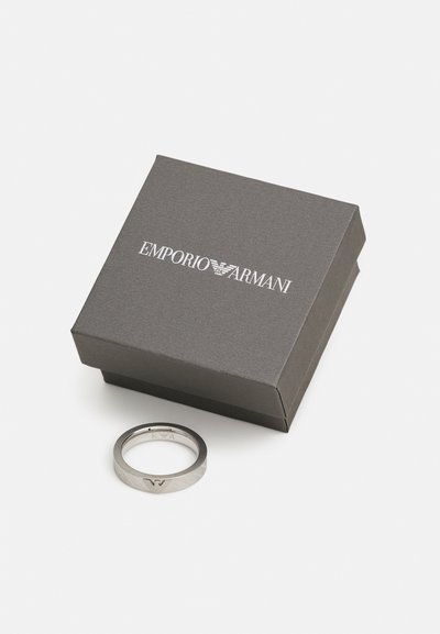 Silver metal ring with a textured surface and engraved logo, placed beside a gray box featuring printed brand name.