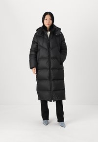 Black puffer coat with a long, hooded design. Features a zip closure, elastic drawstrings, and quilted pattern. Worn over black pants and blue shoes.