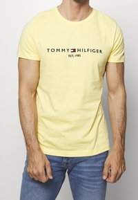 T-shirt en coton jaune avec un col rond, arborant le logo "TOMMY HILFIGER" en noir, rouge et bleu, ainsi que le texte "EST. 1985".