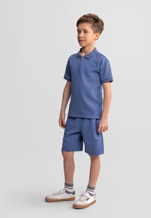 Poiss seisab sinise lühikese varrukaga lukuga polo ja sobivate lühikeste pükstega, triibuliste sokkidega ja valgete tossudega tavalise tausta ees.