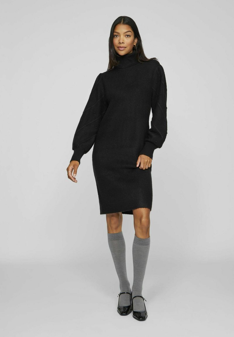 Robe noire en tricot avec un col haut, des manches ballons et des poignets côtelés. Portée avec des chaussettes grises montantes et des chaussures noires à brides.