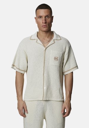 Man draagt een crèmekleurig, gestructureerd kortermig overhemd en bijpassende broek, met beige bies en een borstzak waarop het logo is geborduurd.