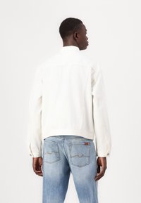 Jachetă din denim alb cu mâneci lungi, manșoane cu buton, și detalii pe spate, asortată cu blugi albaștri deschiși cu accente cusute la buzunare.