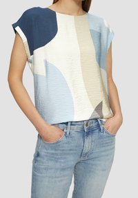 Kortærmet top i lyseblå, creme og mørkeblå med abstrakt mønster; tekstureret stof; kombineret med lyseblå jeans.