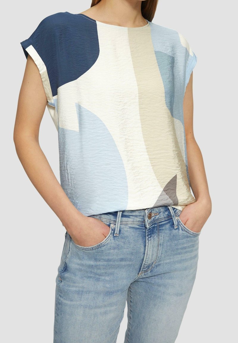 Kortærmet top i lyseblå, creme og mørkeblå med abstrakt mønster; tekstureret stof; kombineret med lyseblå jeans.