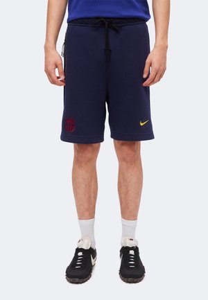 Homme portant un short de sport bleu marine avec cordon noir, logo rouge du FC Barcelone, virgule jaune Nike, chaussettes blanches et baskets Nike noires.
