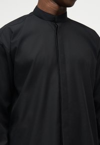 Άνδρας φορώντας μαύρο πουκάμισο με μανδύα μανταρίνου και κρυφά κουμπιά μπροστά, σε απλό φόντο.