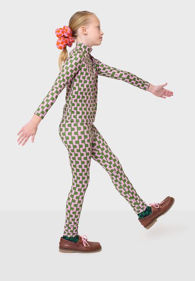 Bunter Langarm-Overall mit einem geometrischen Muster in Grün und Pink, kombiniert mit braunen Schuhen und einem leuchtend orangefarbenen Haargummi.