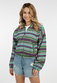 Groene en meerkleurige zip-up jas met patronen van lijnen en bladeren, met een cropped ontwerp en elastische tailleband, gedragen over denim shorts.