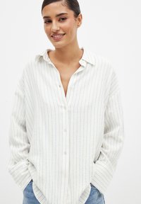 Camicia bianca con righe verticali azzurro chiaro, maniche lunghe, chiusura con bottoni frontale, texture morbida e vestibilità rilassata. Presenta un colletto classico.