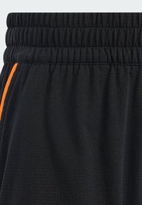 Black athletic shorts featuring an elastic waistband, textured fabric, and an orange side stripe accent. Close-up view of material and design.