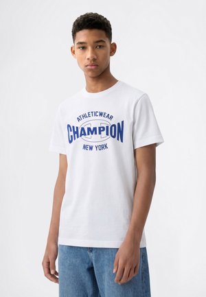 Jeune homme portant un t-shirt blanc avec un texte bleu "Champion Athleticwear New York" et un jean bleu sur un fond clair uni.