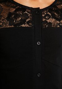 Haut noir à boutons avec empiècement en dentelle florale laissant apparaître la peau autour de l'encolure et du haut de la poitrine.