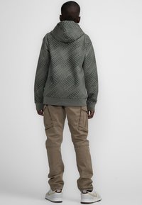 Felpa con cappuccio verde a motivi, con un design testurizzato, abbinata a pantaloni cargo marrone chiaro e sneaker bianche con dettagli dorati.