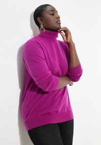 Fuchsia Rollkragenpullover aus weichem Strick mit gerippten Bündchen und Saum. Das Modell posiert mit verschränkten Armen und trägt große kreisförmige Ohrringe. Schwarze Hose.