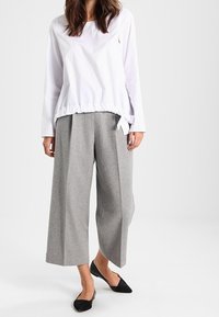 Haut blanc à manches longues avec un cordon de serrage à l'ourlet et un pantalon gris à jambes larges, associé à des ballerines noires, sur un fond uni.