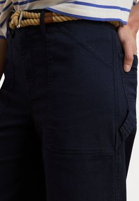 Navy cargobukser laget av bomullsblanding med lomme detaljer. Båret med et beige taubelte og en stripete topp. Nærbilde av stoffets tekstur.