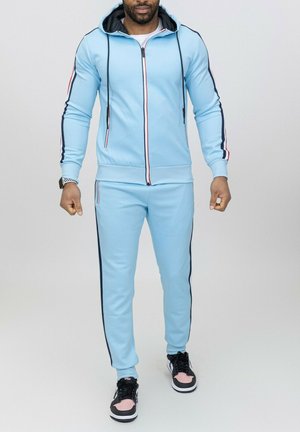 Survêtement bleu clair avec capuche, fermeture éclair sur le devant, présentant des rayures rouges et bleu marine le long des manches et sur les côtés. Porté avec des baskets noires et roses.