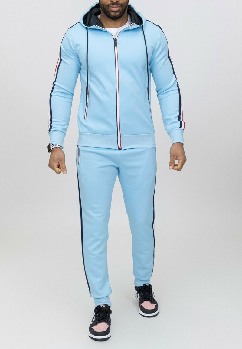 Survêtement bleu clair avec capuche, fermeture éclair sur le devant, présentant des rayures rouges et bleu marine le long des manches et sur les côtés. Porté avec des baskets noires et roses.