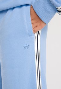 Käsi valgusiniste spordipükside taskus, millel on valged ja mustad külgredelid ning väike embosseeritud logo tasku lähedal.