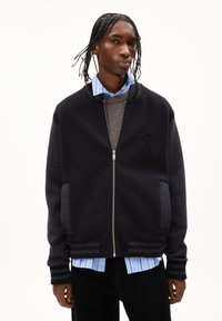 Un jeune homme avec des cheveux tressés portant une veste universitaire zippée noire par-dessus un pull marron et une chemise bleue rayée, se tenant devant un fond blanc.