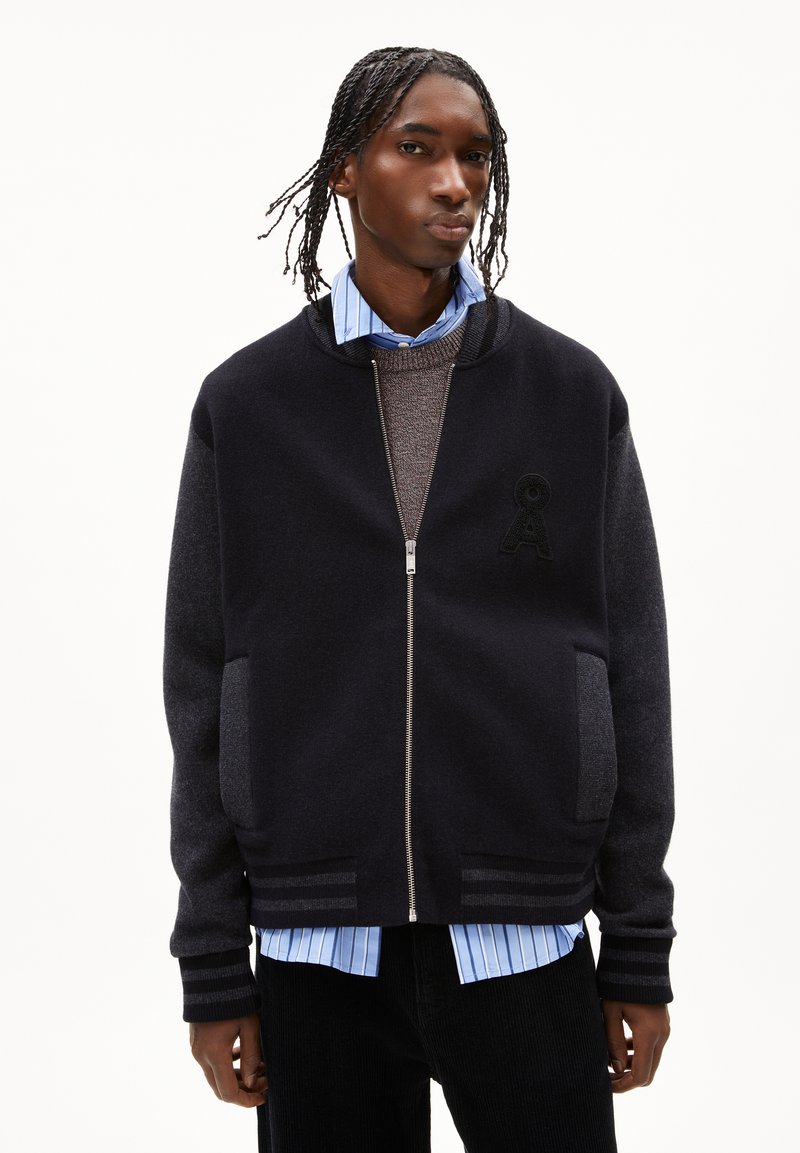 Un jeune homme avec des cheveux tressés portant une veste universitaire zippée noire par-dessus un pull marron et une chemise bleue rayée, se tenant devant un fond blanc.