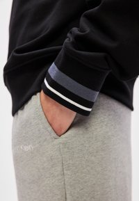 Musta collegepaita, jossa on sinivalkoiset raidalliset hihansuut, yhdistettynä vaaleanharmaisiin collegehousuihin, joissa on hienovarainen logo. Käsi on housujen taskussa.