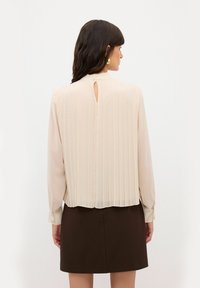 Blusa beige a maniche lunghe con pieghe verticali, dettaglio a chiave sul retro e chiusura con bottone. Abbinata a una gonna marrone scuro.