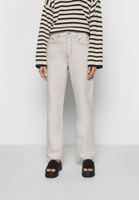 Jeans de cintura alta bege claro com perna reta, costura em contraste, bolsos frontais e bainha ligeiramente desfiada. Combinados com um suéter riscado em preto e branco.
