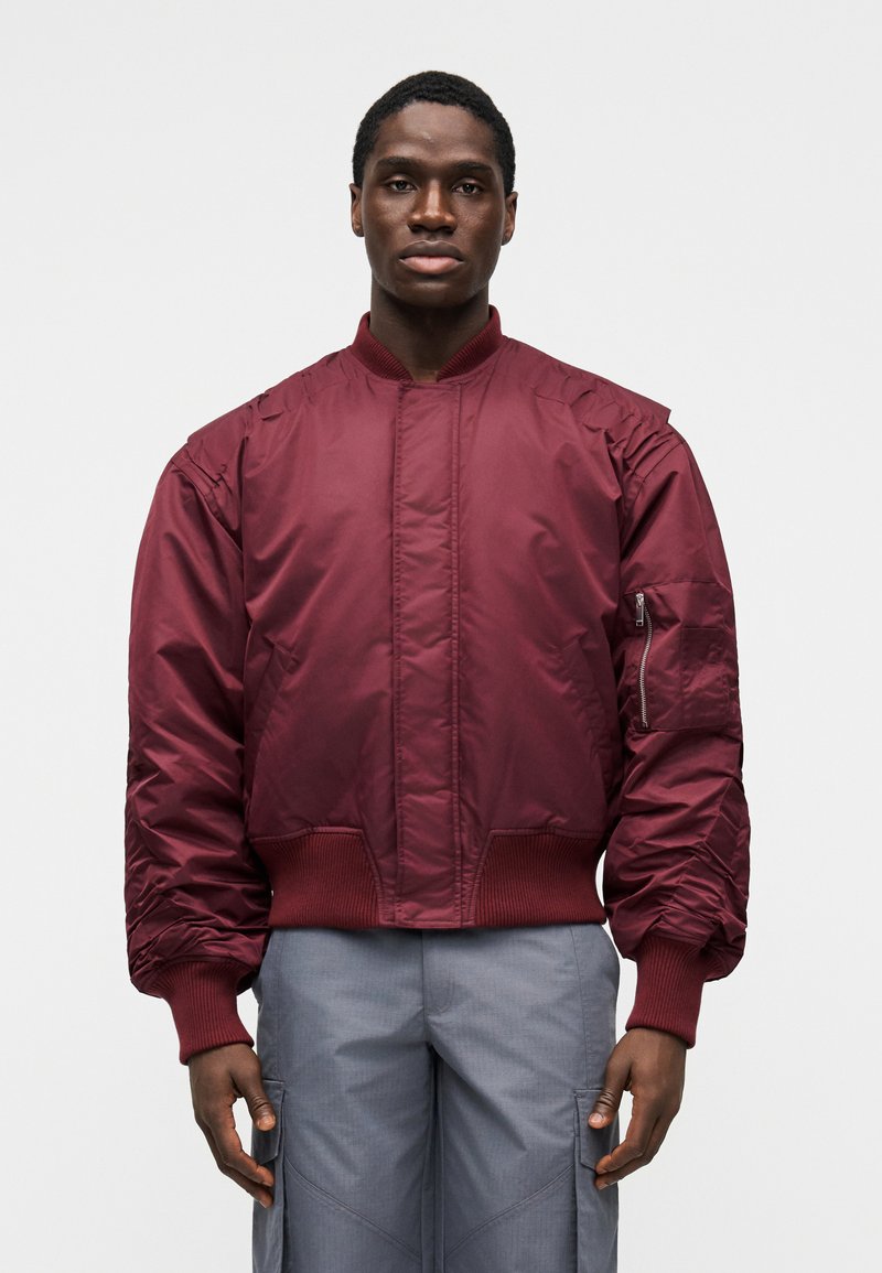 Vínová bomber bunda s rebrovanými manžetami a lemom, s predným vreckom na zips a prešívaným dizajnom pre teplo. Hladká, lesklá textúra látky.