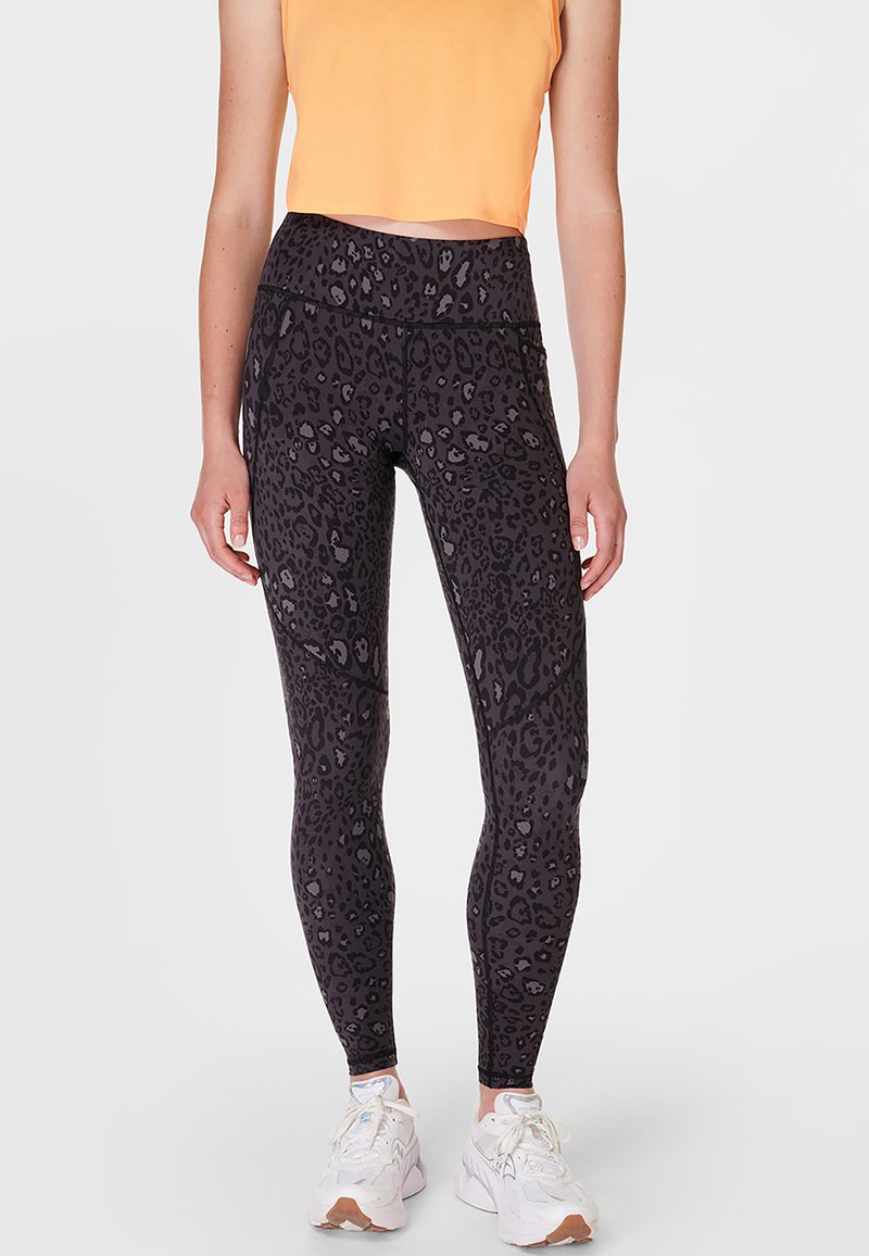 Vrouw in zwarte leggings met luipaardprint, oranje cropped top en witte sneakers, staand tegen een effen lichte achtergrond.