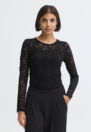 Femme aux cheveux courts et foncés portant un haut noir en dentelle florale à manches longues et un pantalon noir taille haute à plis, main dans la poche.