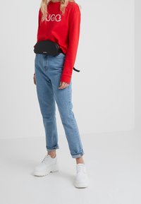 Camisa roja de manga larga con el logo "HUGO", vaqueros de mezclilla azul claro, riñonera negra y zapatillas gruesas blancas. Diseño simple y corte casual.