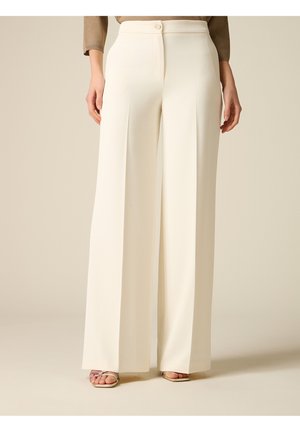 Frau trägt hoch taillierte, weit geschnittene Cremehose mit Vorderfalte, kombiniert mit offenen Sandalen und einem beigen Langarmoberteil.