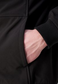 Zwarte jas met een gladde textuur en een zijzak. Een hand rust in de zak, waarbij een gedempte huidtint en zichtbare aderen te zien zijn.