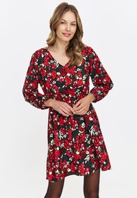 Kobieta z falującymi blond włosami, ubrana w długą sukienkę w kwiaty w kolorze czerwonym, czarnym i białym, stoi na tle jednolitej białej ściany.