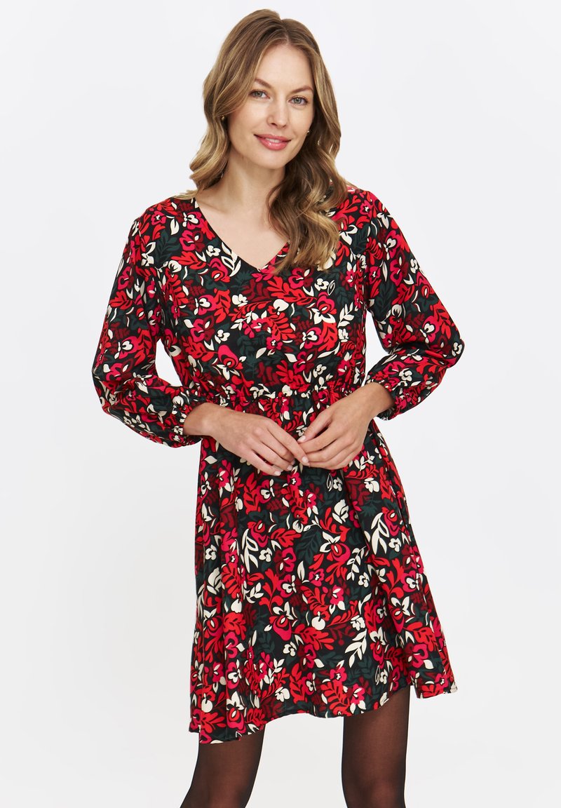 Kobieta z falującymi blond włosami, ubrana w długą sukienkę w kwiaty w kolorze czerwonym, czarnym i białym, stoi na tle jednolitej białej ściany.