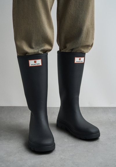 Μαύρες λαστιχένιες μπότες βροχής με στρογγυλερή μύτη, χαμηλό τακούνι, πλευρικές εγκοπές για εύκολη εφαρμογή και ορθογώνια ετικέτα λογότυπου σε λευκό και κόκκινο.