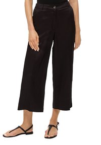 Černé kalhoty s širokými nohavicemi a hladkým povrchem, s knoflíkovým zapínáním a přední kapsou, doplněné černými plochými sandály.