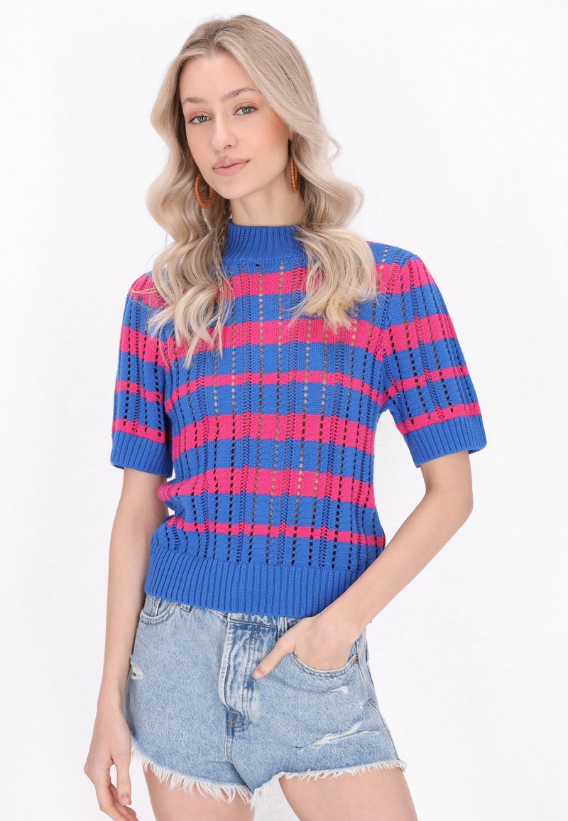 Niebieski i różowy sweter dzianinowy z krótkim rękawem, z poziomym wzorem w paski oraz ściągaczami u rękawów i u dołu, noszony z postarzanymi szortami dżinsowymi.