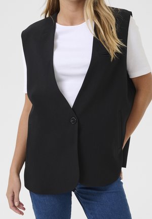 Femme portant un blazer noir sans manches sur un top blanc à manches courtes et un jean bleu, une main dans la poche sur fond blanc.