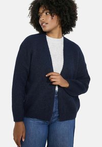 Tums tumši jūras zila virsjakas ar atvērtu priekšpusi, izgatavota no mīkstas, teksturētas adīšanas. Izvēlēta ar platiem piedurknēm un brīvu piegriezumu, kopā ar gaiši pelēku blūzi un zilām džinsām.