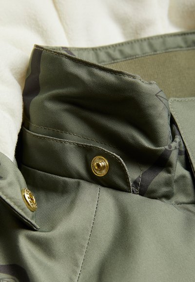 Alyvuogių žalumo marškinių apykaklė su auksiniais mygtuko akcentais ir tekstūruota medžiaga. Turi lygią faktūrą ir švelnų blizgesį.