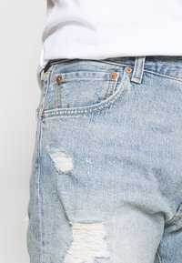 Jeans de mezclilla azul claro con efecto desgastado y orillas deshilachadas, que cuentan con bolsillos y tachuelas de cobre. El material tiene un acabado texturizado con costuras visibles.