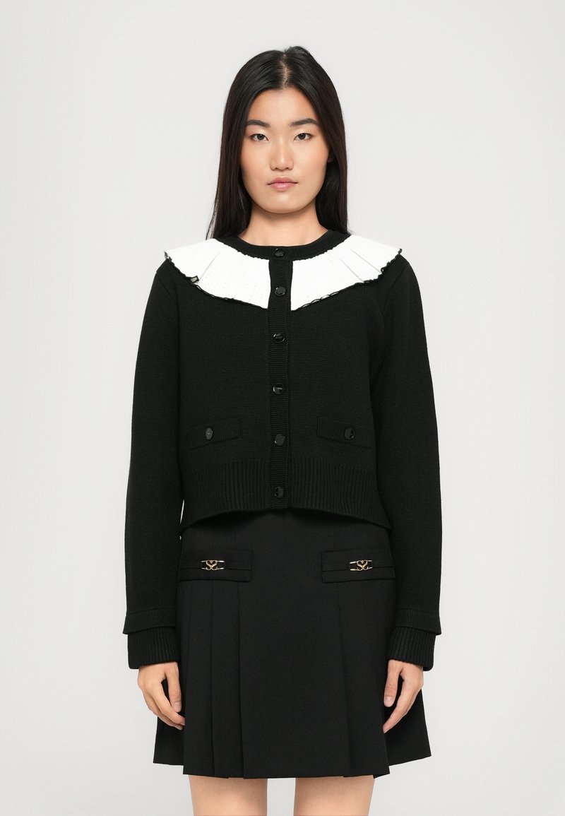 Cardigan nero lavorato a maglia con colletto plissettato bianco, dotato di bottoni frontali e tasche laterali. Indossato sopra una gonna nera coordinata con dettagli.