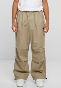 Pantalons cargo beige avec une taille élastique, des poches latérales et une coupe ample. Le matériau est léger avec une texture lisse, accentué par des accessoires noirs.