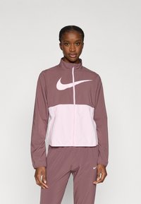 Μπουφάν Nike σε απαλό μωβ και ανοιχτό ροζ, με ψηλό γιακά, πλήρες φερμουάρ και μεγάλο λογότυπο στο στήθος. Λείο, ελαφρύ ύφασμα.