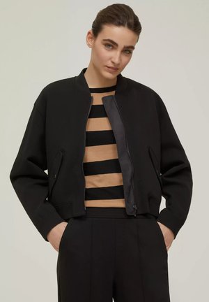 Giacca bomber corta nera con una texture liscia, colletto a coste e tasche laterali. Indossata sopra un top rigato beige e nero.