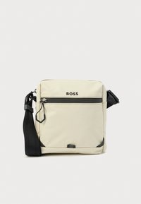 Beež värvi õlakott vastupidavast kangast, ees lukk-tasku, reguleeritav rihm ja mustad detailid. Ees kuvatakse logo.